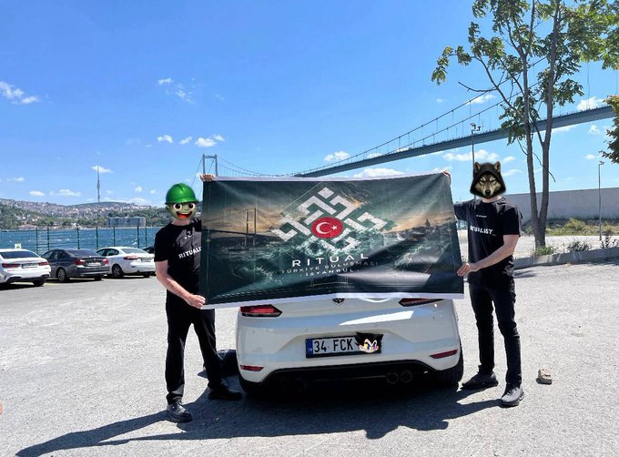 Two people wearing masks (a green hard hat and a wolf mask) stand behind a white car holding a black 'Ritual Uçhisar Kaya Otel' flag with a Turkish crescent and star logo, in front of a bridge over water.