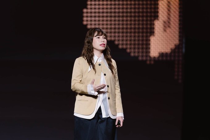 A woman in a tan blazer stands on stage gesturing as she speaks, set against a dark backdrop with an illuminated dot-pattern graphic. The image captures Ms. Nguyễn Minh Hương, CEO of SCEX, delivering her keynote presentation at the Conviction 2026 event.