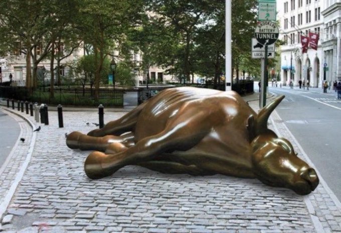 A bronze bull sculpture lying on its side on a cobblestone street in a city, with a 'Battery Tunnel' sign visible nearby.