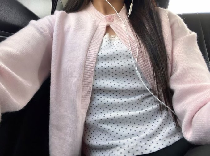 A person wearing a pink cardigan over a white polka-dot top, with white earbuds visible, seated inside a vehicle. Long dark hair falls over one shoulder, and the framing focuses on the torso and neckline.