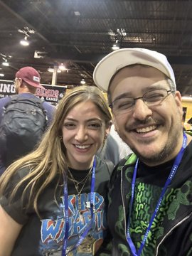 A selfie of a smiling woman and man wearing glasses and a white cap, taken at what appears to be a convention with lanyards around their necks.