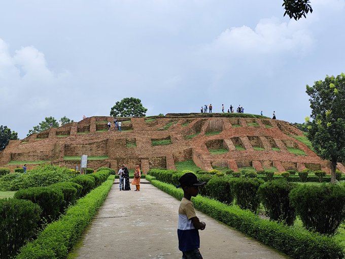 A young boy in a cap walks along a path lined with manicured hedges, with ancient brick ruins and visitors exploring the historic archaeological site in the background, suggesting the poster is sharing a scenic view of a cultural landmark while seeking morning greetings after recovering from illness.
