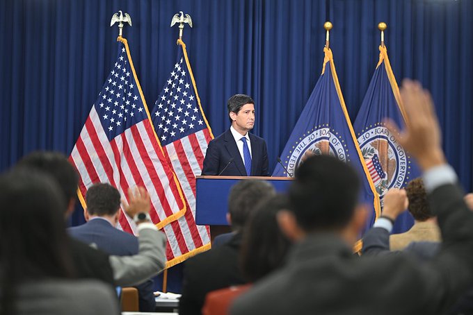 A man in a suit speaks at a podium flanked by American flags and U.S. Department of Justice flags, addressing an audience of reporters with raised hands.