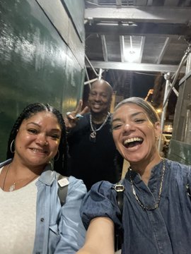 Three people pose for a selfie on a dark street at night, with two smiling women in the foreground and a man making hand gestures behind them.