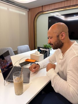 A bald man with a beard wearing a white long-sleeve shirt sitting at a desk with a laptop and a glass of a light-colored beverage.