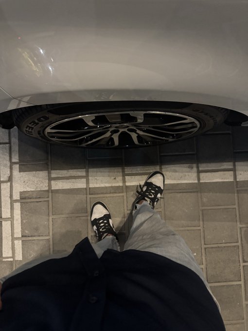 A first-person perspective photo looking down at a person's feet in black and white sneakers standing on a tiled floor next to a white car with alloy wheels.