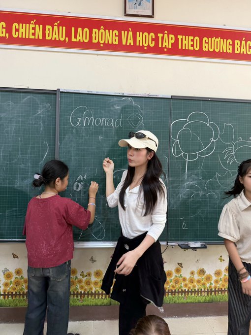A woman wearing a beige cap and white shirt draws on a chalkboard next to a young girl, with "Gmonad" written in chalk and a Vietnamese banner reading "...G, CHIẾN ĐẤU, LAO ĐỘNG VÀ HỌC TẬP THEO GƯƠNG BÁC" displayed above. The image appears to capture a creative classroom scene promoting the "Gmonad" project, blending educational graffiti art with an inspiring Vietnamese motto about striving, working, and studying as an example.