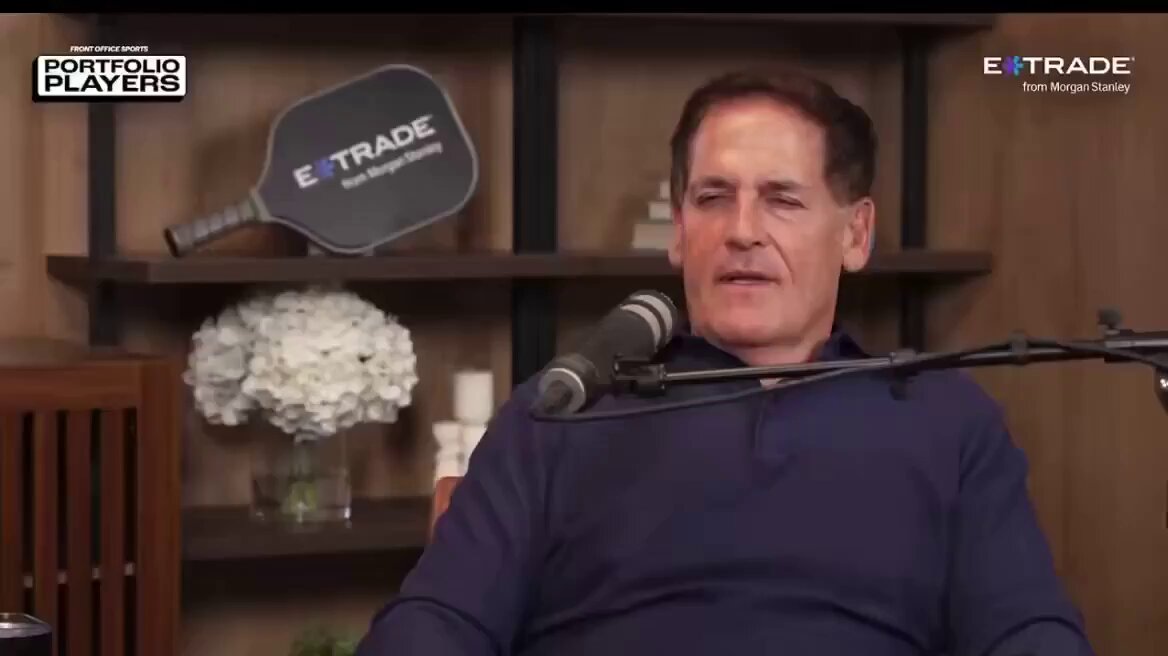 The image features a man wearing a blue shirt, sitting down with a microphone in front of him. He appears to be talking about Bitcoin or cryptocurrency, as indicated by the text "Mark Cuban on Bitcoin: I’ve sold most of it." The man is seated at a desk and there are two potted plants nearby - one located close to the camera and another further away.
