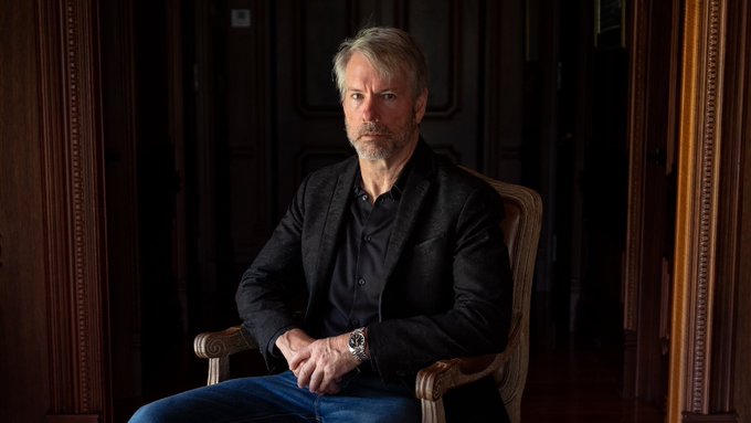 A bearded man in a black blazer sits contemplatively in an ornately carved wooden chair within a dimly lit, wood-paneled room, his expression serious as he gazes off-camera. The accompanying caption ties this portrait to Michael Saylor, who has warned that internal governance shifts within Bitcoin—not outside rivals—pose the greatest risk to the cryptocurrency's scarcity and long-term security.