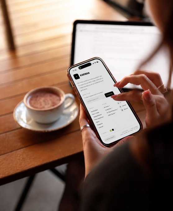 An over-the-shoulder view of a person using a smartphone to fill out an online application form for 'KARVAN' while sitting at a wooden table with a coffee cup and laptop.