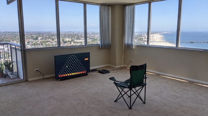 A room with floor-to-ceiling windows offering a panoramic ocean view, featuring a green folding chair and a TV displaying a colorful geometric pattern.