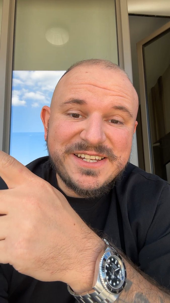 A smiling, bald man with a goatee and mustache takes a selfie indoors near a window showing blue sky, wearing a black shirt and a Rolex watch on his wrist.