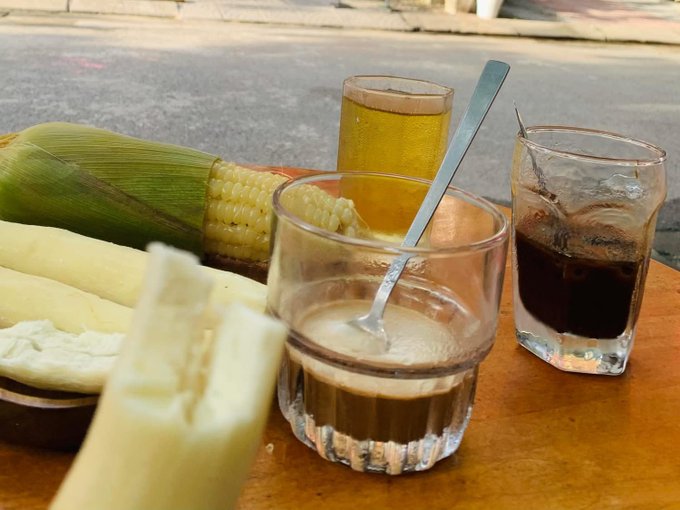 A street-side table displays glasses of coffee and tea alongside a plate of corn and white cheese, likely a traditional South American breakfast.