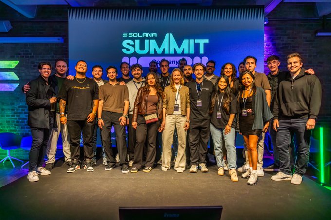 The image features a group of people standing together on stage at an event called Solana Summit. The attendees are posing for a photo with smiles on their faces, showcasing their enthusiasm and camaraderie. The backdrop includes a large screen displaying the text "Solana Summit" in blue letters, indicating that this is a gathering related to the Solana cryptocurrency project or its summit.