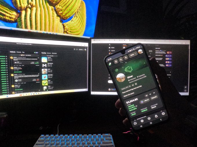 A person holds a smartphone displaying a crypto wallet or trading app in front of two computer monitors showing similar interfaces.