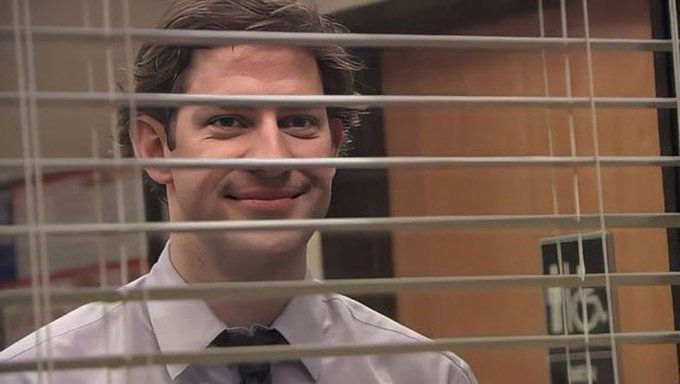 A smiling man in a white shirt and tie looking through window blinds, recognizable as Jim Halpert from The Office.