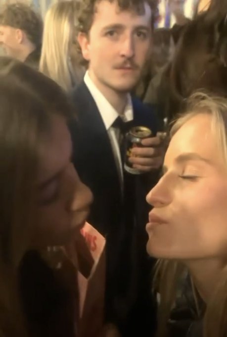 The image shows two women in the foreground standing very close together with their faces near each other, while a young man in a suit and tie stands behind them holding a beverage can, staring blankly ahead as if oblivious to the women right in front of him—fitting the meme's humor about crypto enthusiasts being out of touch in social situations.