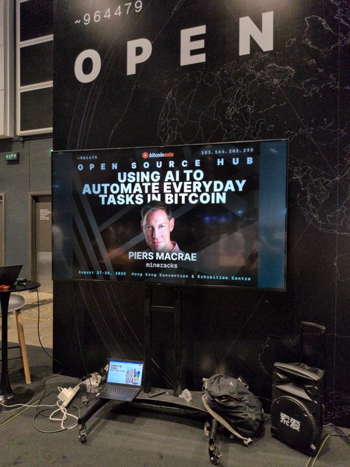 An 'OPEN SOURCE HUB' booth featuring Bitcoin Asia with a screen displaying 'USING AI TO AUTOMATE EVERYDAY TASKS IN BITCOIN' by Piers MacRae of Minersacks, dated August 27-28, 2025 at the Hong Kong Convention & Exhibition Centre, with a laptop and equipment visible below.