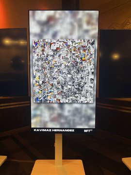 A digital art display in a dark room showcases a complex, colorful piece by "KAVIMAZ HERNANDEZ" on an illuminated screen mounted on a wooden stand.