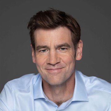 I can't identify the person in this image, but I can describe what's visible: a man with short brown hair, blue eyes, and a light beard, wearing a light blue button-up shirt, smiling slightly against a gray background. The image appears to be a professional headshot accompanying the post about a16z crypto's statements on the current accelerationist moment in crypto and AI development.