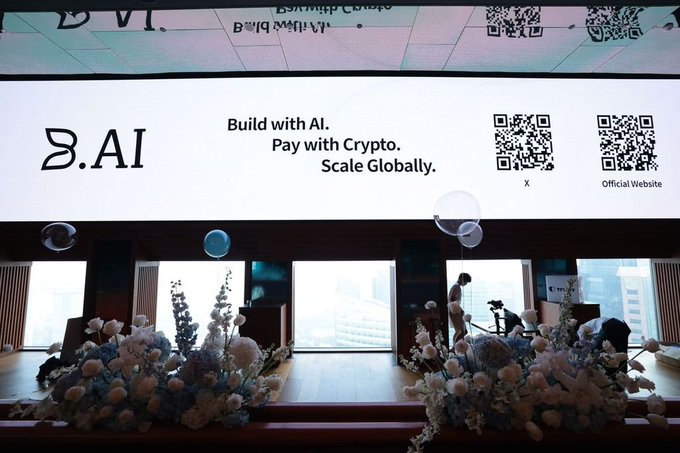 An indoor event space with floral arrangements in the foreground and a large screen displaying '.AI' with the text 'Build with AI. Pay with Crypto. Scale Globally.' along with QR codes for X and the Official Website.