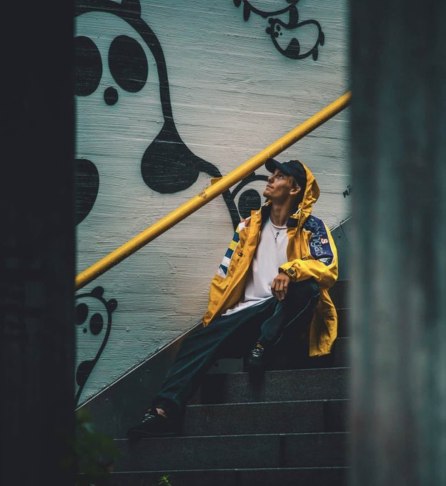 A young man wearing a yellow hooded rain jacket sits on outdoor concrete steps, looking up against a wall decorated with cartoonish black graffiti.