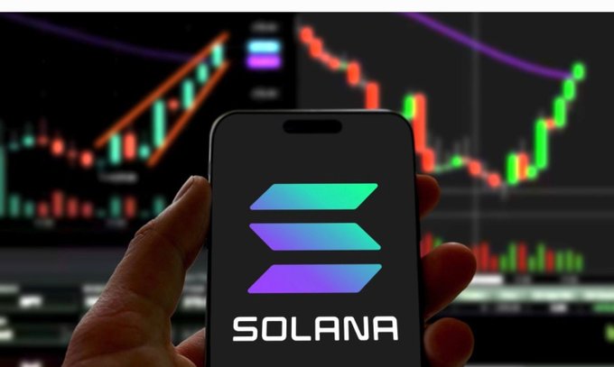 A hand holding a smartphone displaying the Solana logo against a background of cryptocurrency trading charts with green and red candlestick patterns.