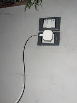 A white phone charger is plugged into a wall outlet against a plain gray wall, with its cable dangling down—capturing the desperate 6 AM scramble to charge a nearly dead phone for the OpenSea beta code, as mentioned in the post.