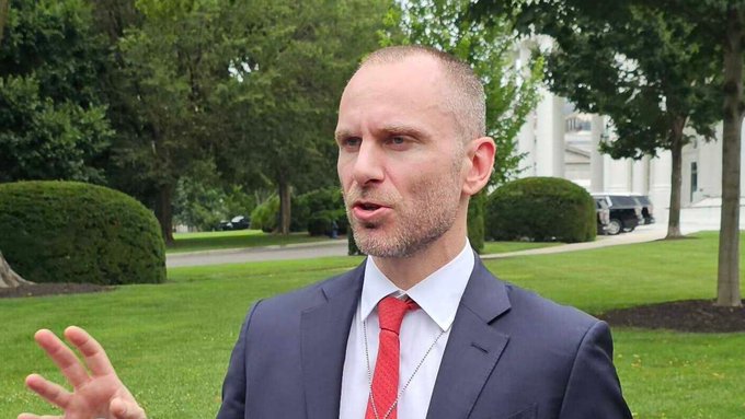 A bald man with a gray beard wearing a dark suit and red tie gestures with his hand while speaking outdoors with green lawns and trees behind him.