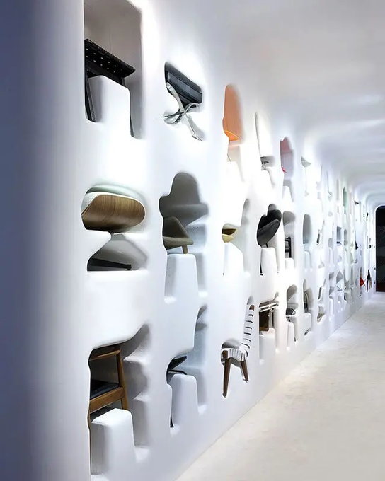 An interior view of a modern furniture showroom featuring a white wall with cutouts displaying various designer chairs and seating.