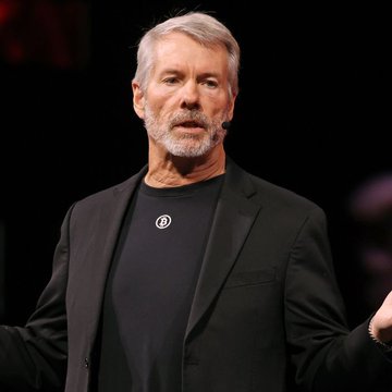 A gray-bearded man in a black blazer and t-shirt featuring a circular logo stands on stage with arms slightly extended, appearing mid-speech. The image likely depicts Michael Saylor, whose commentary about Bitcoin purchases ("Orange dots tell only part of the story") accompanies the post discussing Strategy's potential Bitcoin sales.