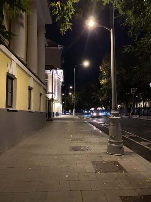 A nighttime view of an empty sidewalk along a city street, illuminated by streetlights, with a yellow building on the left and parked cars on the right.