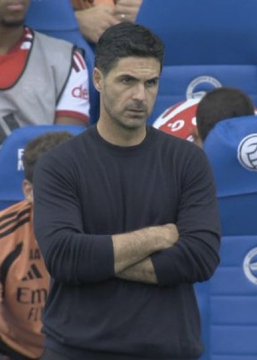 A photograph of Arsenal manager Mikel Arteta standing on the sidelines with his arms crossed, wearing a dark navy sweater.