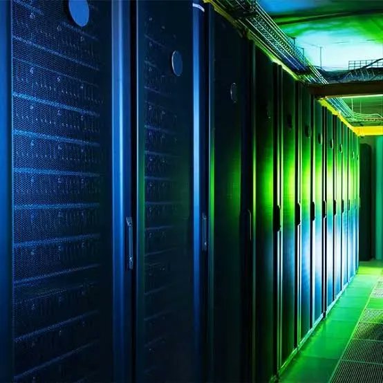 Rows of black server racks illuminated by blue and green LED lights line a corridor in a data center, visually representing the massive physical infrastructure required to support the projected $2.7 trillion in AI spending by 2026. The endless rows of compute hardware underscore the post's point that AI's exponential growth is fundamentally bottlenecked by who controls and builds these critical data center facilities.