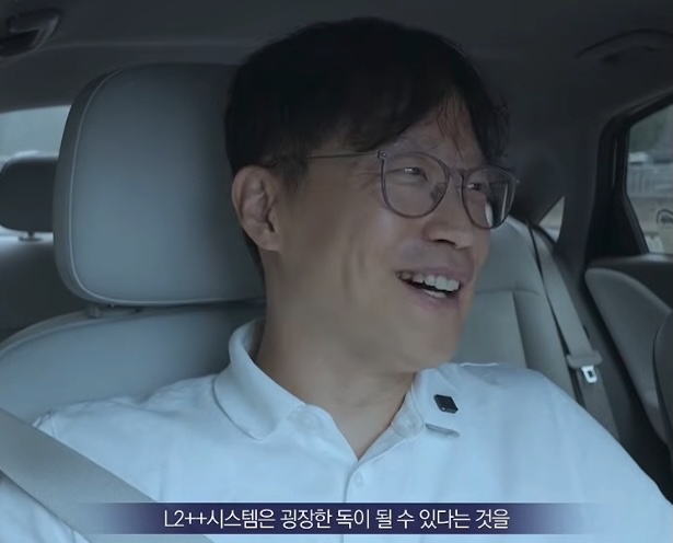 A man wearing glasses and a white polo shirt is seated in the driver's seat of a car, smiling broadly with a seatbelt visible across his chest. Korean subtitles at the bottom of the image read "L2++시스템은 굉장한 독이 될 수 있다는 것을," which translates to "An L2++ system can become a tremendous poison," visually accompanying CEO Park Min-woo's warning about the dangers of over-reliance on advanced driver-assistance technology.