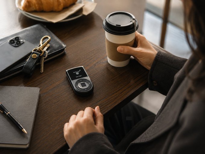 A person sits at a wooden café table holding a takeaway coffee cup, with their keys, smartphone, and a smart key tracker device placed nearby. The image visually reinforces the post's message by showing how the key finder fits seamlessly into a daily routine, keeping essentials secure alongside morning coffee and personal items.