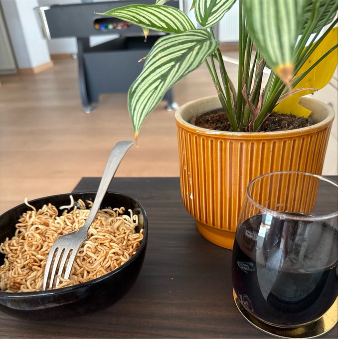 A bowl of instant noodles with a fork sits on a dark wooden table next to a potted calathea plant and a glass of dark liquid.