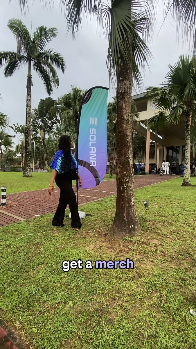 A woman in a blue shirt and black pants walks past a tall, blue and purple "SOLANA" feather flag at an outdoor venue with palm trees, with text overlay reading "get a merch".