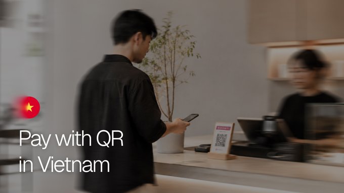 A man holds his phone to scan a QR code on a counter at a Vietnamese shop, with on-screen text reading "Pay with QR in Vietnam." The image visually illustrates the post's message about enabling tourists to pay with crypto via Vietnam's widely-used QR payment system.