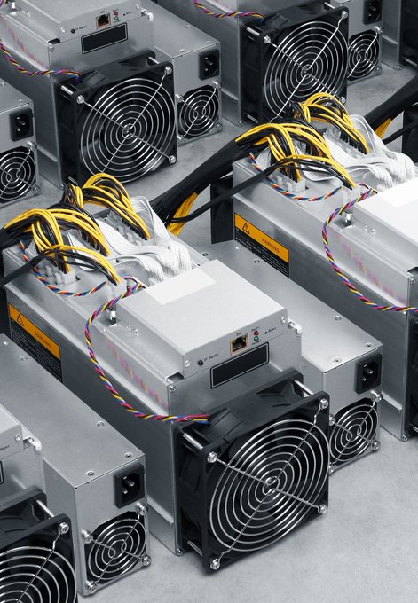 A row of cryptocurrency mining rigs with cooling fans and colorful wiring sits on a warehouse floor, illustrating the physical hardware behind Bitcoin mining operations. The image accompanies news of former leading Bitcoin miner POOLIN's Chapter 11 filing, depicting the type of equipment that contributed to its rise to an 18% network hashrate before its collapse due to excessive financial leverage.