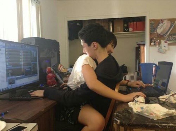 Two people sit back-to-back at a cluttered desk with two computer monitors, appearing to use the computers simultaneously.