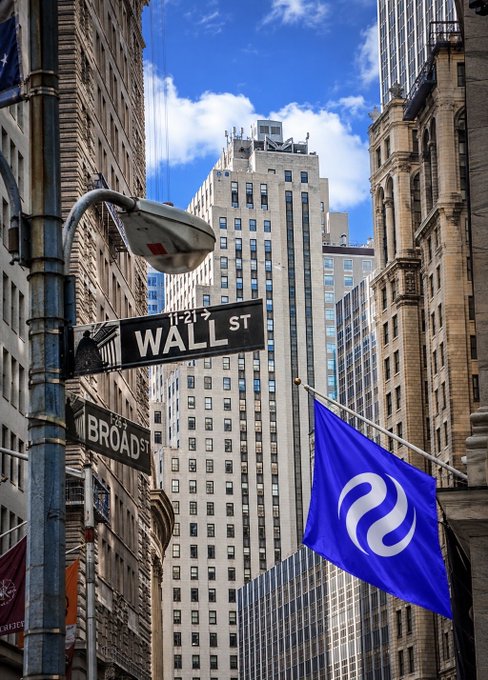 A street view at the intersection of Wall St and Broad St in New York City's financial district, marked by classic green street signs and a blue flag with a white swirling logo, symbolizing a token or cryptocurrency's arrival to Wall Street as referenced by the "$INJ" ticker in the post.