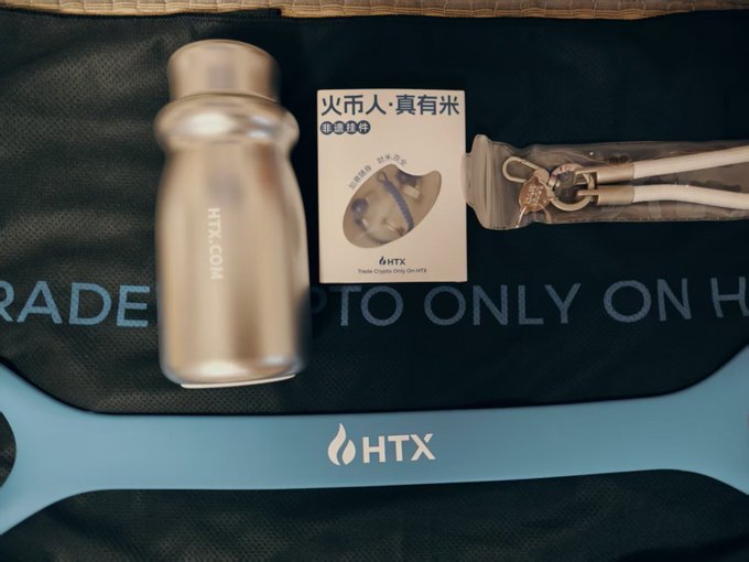 A flat lay photo showing HTX-branded merchandise including a silver HTX.COM water bottle, a card featuring a Shiba Inu with Chinese text "火币人·真有米" (HTX People Really Have Rice) advertising "6mHTX Trade Crypto Only On HTX", an umbrella in a pouch, and a blue HTX-branded item, all arranged on a black bag with "CRYPTO ONLY ON H..." text visible.