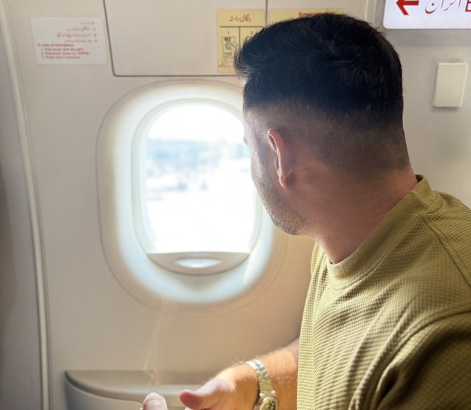 A man with a short fade haircut looks out the oval window of an airplane during the day.