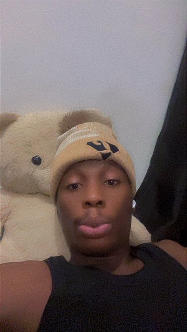 A person wearing a black tank top and a yellow beanie takes a selfie next to a large cream-colored teddy bear.