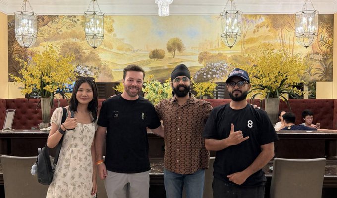 Four people stand side-by-side smiling at the camera in a restaurant with decorative pendant lighting and a landscape mural on the wall behind them.