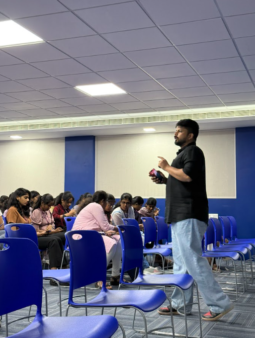 A man in a black shirt stands holding a microphone, addressing a seated audience in a blue conference room.