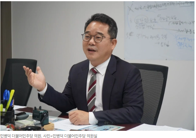A middle-aged Korean man in a suit and glasses gesturing while speaking at a desk, with a whiteboard behind him covered in writing; the caption reads "민병덕 더불어민주당 의원, 사진=민병덕 더불어민주당 의원실" (Min Byung-duk, Democratic Party lawmaker, photo=Min Byung-duk's office).