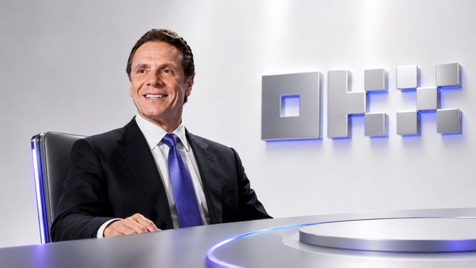 A portrait of a smiling man in a dark suit and blue tie seated in front of a DHX logo display.
