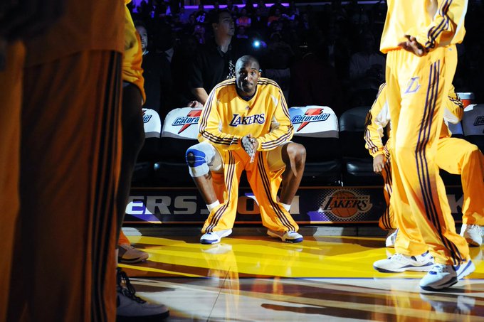 A Lakers basketball player in a yellow warm-up suit crouching on the court during pregame, with other players visible nearby.